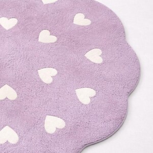 May include: A lavender-colored, heart-shaped rug with a scalloped edge. The rug is covered in white heart-shaped designs. The rug has a soft, plush texture, and is likely made of a synthetic material.
