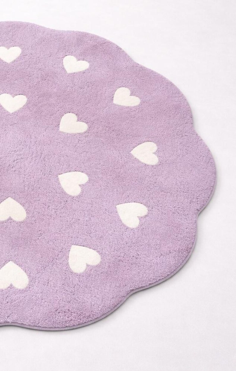 May include: A lavender-colored, heart-shaped rug with a scalloped edge. The rug is covered in white heart-shaped designs. The rug has a soft, plush texture, and is likely made of a synthetic material.