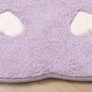 May include: A soft, lavender-colored rug with a scalloped edge and a repeating pattern of white hearts. The rug features a plush, textured pile surface and is finished with a matching lavender fabric trim along the curved border.
