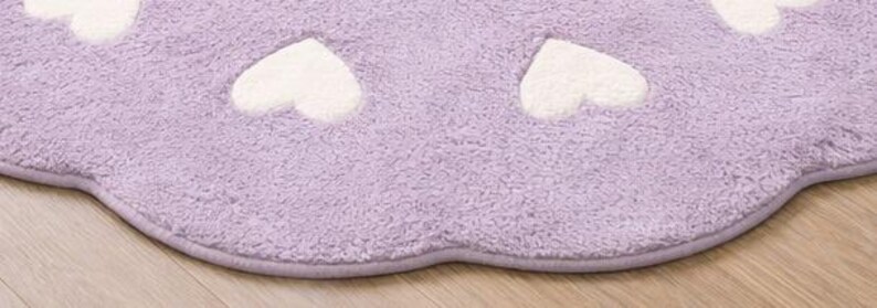 May include: A soft, lavender-colored rug with a scalloped edge and a repeating pattern of white hearts. The rug features a plush, textured pile surface and is finished with a matching lavender fabric trim along the curved border.