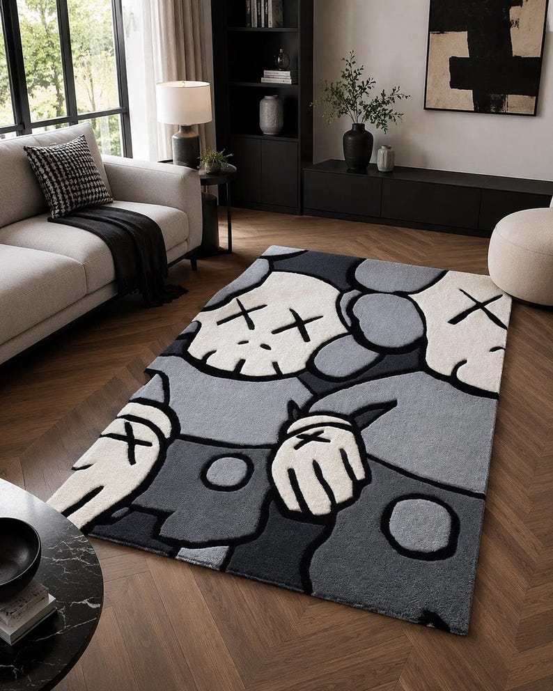 Puede incluir: Una alfombra rectangular con un dise&ntilde;o de personaje de dibujos animados estilizado en tonos gris, blanco y negro. El personaje tiene una cabeza similar a una calavera con ojos en forma de X, manos enguantadas y una superficie texturizada.