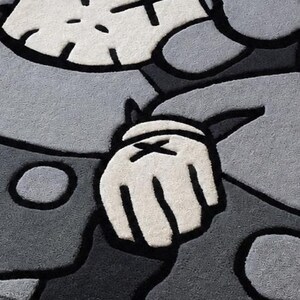 Puede incluir: Una alfombra de pelo cortado con un dise&ntilde;o de personaje estilizado en tonos gris claro, gris oscuro y blanco. El gr&aacute;fico incluye una cabeza similar a una calavera con ojos en forma de X y una mano enguantada con una X, delineado en negro.