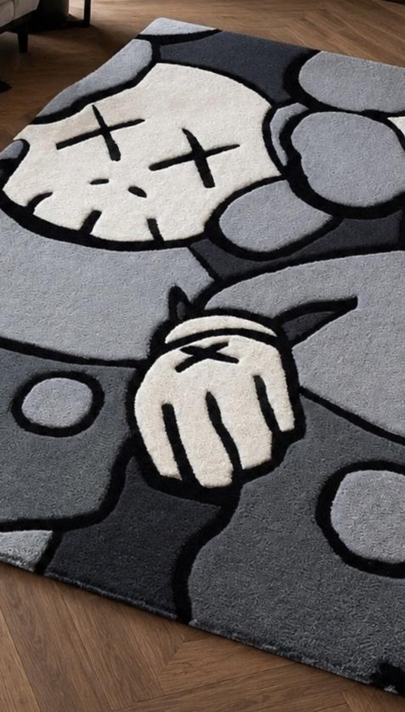 Puede incluir: Una alfombra de pelo cortado con un dise&ntilde;o de personaje estilizado en tonos gris claro, gris oscuro y blanco. El gr&aacute;fico incluye una cabeza similar a una calavera con ojos en forma de X y una mano enguantada con una X, delineado en negro.