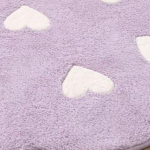 May include: A close-up of a lavender-colored, plush rug with white heart-shaped accents. The rug has a soft, textured appearance, with the hearts evenly spaced across the surface. The rug's edges are scalloped.