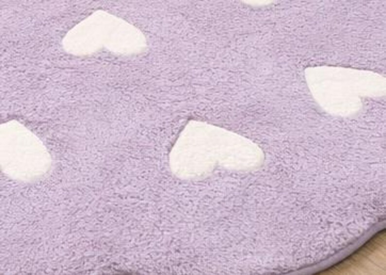 May include: A close-up of a lavender-colored, plush rug with white heart-shaped accents. The rug has a soft, textured appearance, with the hearts evenly spaced across the surface. The rug's edges are scalloped.