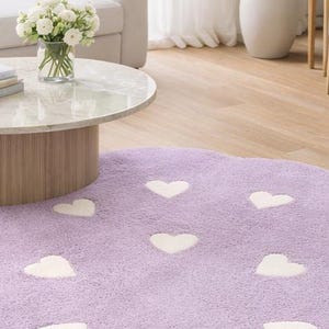 May include: A lavender area rug with a scalloped edge and a repeating pattern of white hearts. The rug has a soft, textured pile and is shaped like a cloud, featuring multiple white heart shapes distributed across the purple surface.
