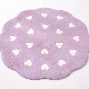 May include: A round, lavender-colored rug with a scalloped edge. The rug features a repeating pattern of small, white heart shapes arranged in a circular formation across the soft, textured surface of the fabric.