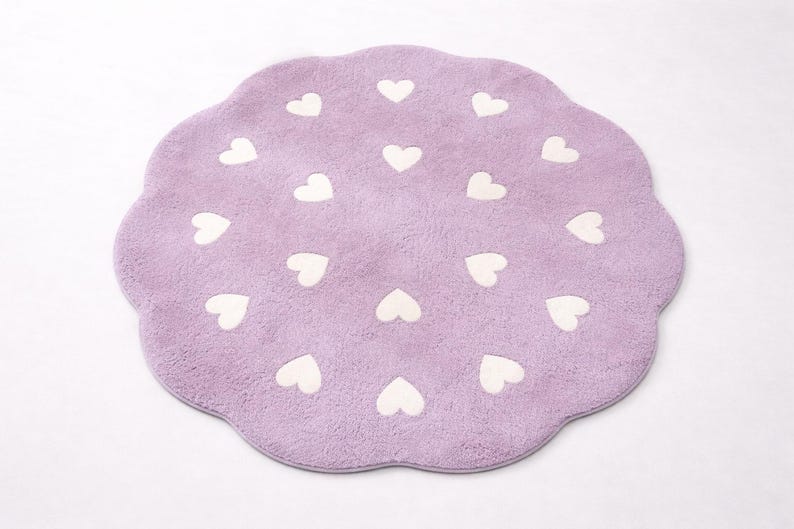 May include: A round, lavender-colored rug with a scalloped edge. The rug features a repeating pattern of small, white heart shapes arranged in a circular formation across the soft, textured surface of the fabric.
