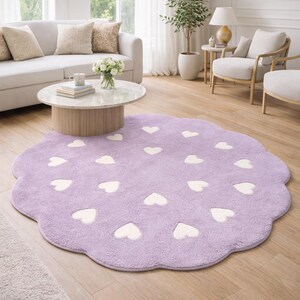May include: A round, scalloped-edge area rug in a soft lavender color, featuring a repeating pattern of small, cream-colored heart shapes distributed across the surface. The rug has a plush, textured pile and a distinct flower-like shape.
