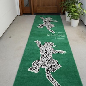 Op de afbeelding: Groen loperkleed met een luipaardprint ontwerp. Het kleed heeft een wit en zwart luipaard silhouet met de tekst "IMPERIAL TUFT AND KNOT" in het wit. Het kleed is geplaatst voor een houten deur.