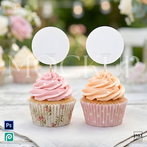 May include: Two cupcakes with pink and peach frosting, topped with white circles on wooden sticks. The cupcakes sit on a white cloth, one in a floral liner, the other in a pink polka dot liner. The background is blurred.