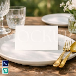 May include: A white rectangular card with the word "MOCKUP" in light gray text, placed on a white plate with gold silverware. The scene includes a glass of water, a wine glass, and a small vase with white flowers. The background is blurred.