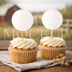 May include: Two cupcakes with white frosting and a blank white circle topper on a wooden stick. The cupcakes sit on a linen napkin on a wooden surface. The background is blurred, showing a barn and field.