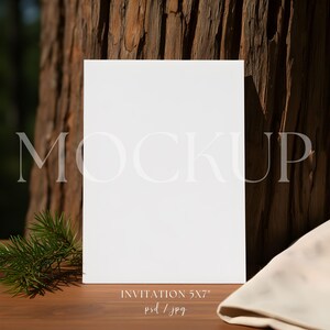 以下が含まれることがあります： 背景に「MOCKUP」という文字が入った、白の12.7 x 17.8 cmの招待状モックアップ。招待状は木の表面に置かれ、松の小枝と折りたたまれた布が添えられています。背景には木の幹があります。