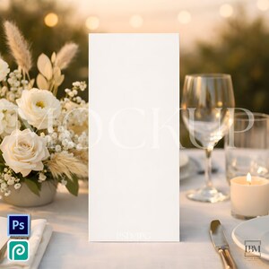 May include: A blank white rectangular card stands upright on a table set for an outdoor event. The table is covered with a white tablecloth and decorated with white flowers, candles, and glassware. The background is blurred with soft lighting.