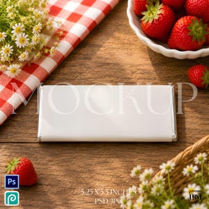 May include: A white rectangular product mockup, 13.3 x 14 cm, on a wooden surface. The scene includes fresh strawberries, daisies, and a red and white checkered cloth. The mockup is wrapped in silver foil.