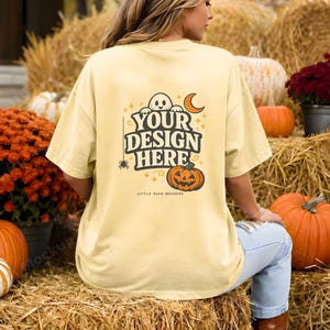 May include: A pale yellow t-shirt featuring a back graphic with a ghost, a crescent moon, a spider, and a jack-o'-lantern pumpkin. The text reads "YOUR DESIGN HERE" in a bold, dark font, with "LITTLE BASH MOCKUPS" printed in smaller text below.
