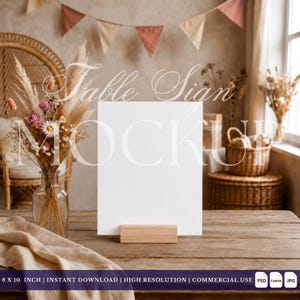 May include: A vertical white rectangular sign mockup held by a light wood block stand. Next to it is a clear glass vase filled with dried pampas grass and pink and white flowers. The text "Table Sign MOCKUP" is overlaid on the image.