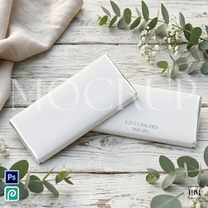 May include: Two white chocolate bar wrappers with silver edges are displayed on a rustic, white wooden surface. The wrappers have the text "MOCKUP" and "5.25 X 5.5 INCHES PSD JPG". Greenery and white flowers surround the bars.