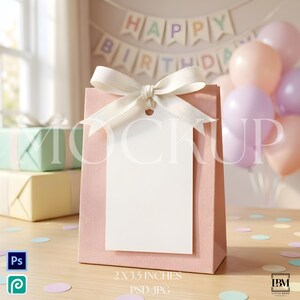 Pode incluir: Um saco de presente de papel rosa claro com uma etiqueta retangular branca presa por um laço de fita cor de creme. O saco está sobre uma superfície de madeira, com um fundo desfocado de balões em tons pastel e uma faixa de Happy Birthday.