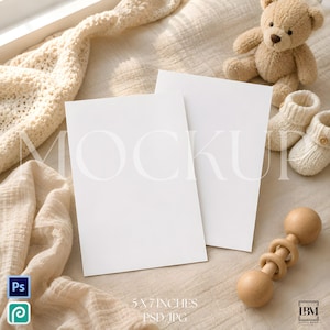 May include: Two blank white rectangular cards measuring 5 by 7 inches, positioned next to a light brown teddy bear, a pair of cream-colored knitted baby booties, and a wooden baby rattle on a soft, textured, light-colored fabric surface.