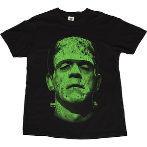 May include: Black t-shirt featuring a green graphic of Frankenstein's monster. The monster's face is prominently displayed, with a textured, distressed effect. The design is centered on the shirt, creating a bold and eye-catching look. A classic horror movie theme.