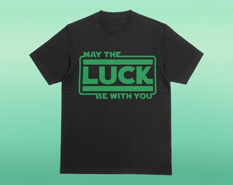 以下が含まれることがあります： 黒いTシャツ。緑色の文字で「MAY THE LUCK BE WITH YOU」と書かれています。フレーズは長方形のボックス内にあり、中央に「LUCK」の文字があります。Tシャツはクルーネックで半袖です。背景は緑色のグラデーションです。