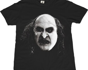 以下が含まれることがあります： 黒いTシャツ。白黒の画像で、肌が白く、髪が黒く、不気味な表情のキャラクターが描かれています。キャラクターの顔が中心で、鼻が大きく、歯が見えています。シャツは定番のクルーネックです。