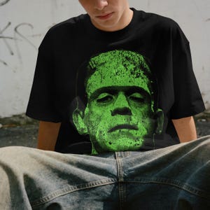 May include: Black t-shirt featuring a green graphic of Frankenstein's monster. The graphic is a close-up of the monster's face, with a textured, distressed appearance. The shirt is a classic crew neck style.