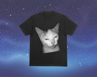 以下が含まれることがあります： 黒いTシャツ。白黒の猫の顔の画像がプリントされています。猫の顔はTシャツの中央に配置され、目は閉じられています。背景は星空です。