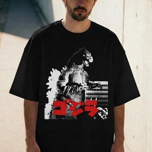May include: Black oversized t-shirt featuring a Godzilla graphic in white and red. The design includes the Japanese word "ゴジラ" in red. The shirt has short sleeves and a relaxed fit, suitable for casual wear.