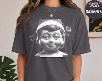 以下が含まれることがあります： グラファイトグレーの半袖Tシャツ。帽子をかぶった人形のグラフィックプリントが施され、頭の周りに煙が渦巻いている。画像の右上隅には「COMFORT COLORS GRAPHITE」というテキストが印刷されている。