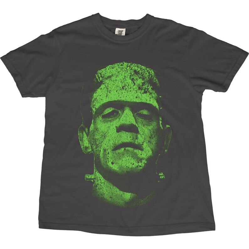 May include: Dark gray t-shirt featuring a distressed green image of Frankenstein's monster. The graphic is a close-up of the monster's face, with visible stitches and bolts. The shirt is a classic crew neck style.