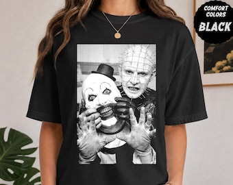May include: A black t-shirt featuring a black and white graphic print of Art the Clown and Pinhead. The design shows Art the Clown wearing a small top hat and Pinhead with pins in their head. The shirt includes a circular black label that reads COMFORT COLORS BLACK.