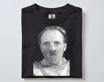以下が含まれることがあります： 黒いTシャツで、口元に金属製のケージが付いた硬いフェイスマスクを着用した人物の白黒写真プリントが施されています。Tシャツはクルーネックで、襟の内側にComfort Colorsのブランドラベルがプリントされています。