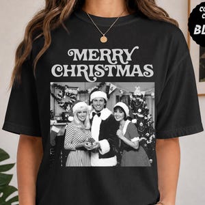 Puede incluir: Una camiseta negra con el texto MERRY CHRISTMAS en una fuente retro blanca sobre una fotografía en blanco y negro de tres personas con gorros de Papá Noel frente a un árbol de Navidad decorado. La camiseta tiene cuello redondo y mangas cortas.