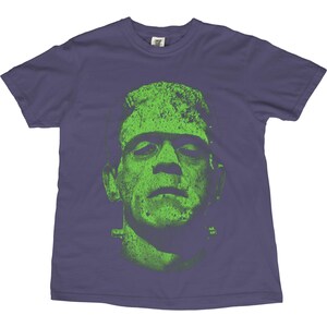 May include: A purple t-shirt featuring a green graphic of Frankenstein's monster. The monster's face is the focal point, with a textured, distressed appearance. The shirt is a classic crew neck style.