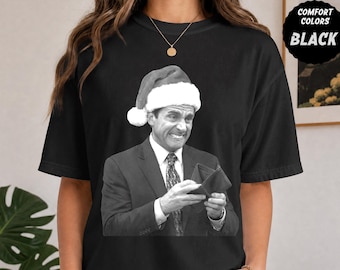 以下が含まれることがあります： 黒いTシャツで、スーツとネクタイを着用し、赤と白のサンタの帽子をかぶって開いた財布を持っている男性のグラフィックプリントが施されています。グラフィックは白黒のトーンです。隅にある黒いバッジには「COMFORT COLORS BLACK」と書かれています。