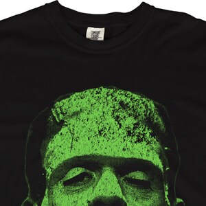 May include: Black t-shirt featuring a green graphic of Frankenstein's monster. The design is a textured, halftone image of the monster's face. The shirt has a crew neck and a tag that says "Comfort Colors".
