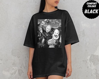 May include: A black t-shirt featuring a black and white graphic print of two horror characters, one holding a bottle and the other a red plastic cup, with a circular badge in the top right corner that reads COMFORT COLORS BLACK.