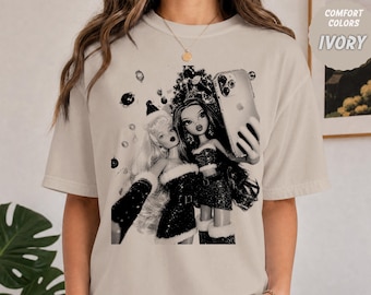 以下が含まれることがあります： アイボリーのTシャツで、サンタ帽をかぶりクリスマス風の衣装を着た2体の人形がスマートフォンで自撮りをしている白黒のグラフィックプリントが施されています。背景にはクリスマスツリーとオーナメントが描かれています。