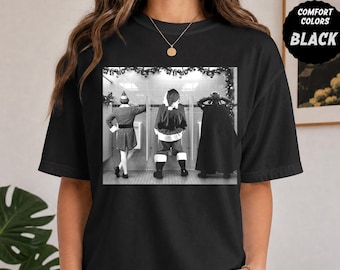 以下が含まれることがあります： 黒いTシャツで、エルフ、サンタクロース、ダース・ベイダーがトイレの小便器の前に立っている白黒のグラフィックプリントが施されています。右上隅には、COMFORT COLORS BLACKというテキストが入った黒い円形のバッジがあります。