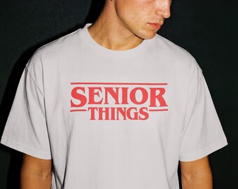 以下が含まれることがあります： 「SENIOR THINGS」の赤い文字がプリントされたライトグレーのTシャツ。テキストは中央に配置され、レトロなデザインです。Tシャツはクルーネックです。