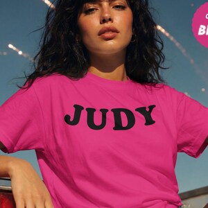 May include: A berry-coloured short-sleeve t-shirt featuring the name JUDY printed in bold black capital letters across the chest. A circular pink badge in the upper right corner displays the text BELLA CANVAS BERRY in white lettering.