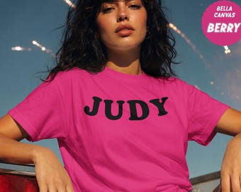 以下が含まれることがあります： 胸元に太い黒の大文字でJUDYとプリントされたベリー色の半袖Tシャツ。右上の円形のピンクのバッジには、白い文字でBELLA CANVAS BERRYと記載されています。