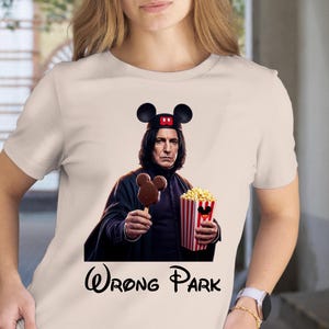 May include: A cream-colored t-shirt featuring a parody image of a character wearing a Mickey Mouse hat and holding a Mickey Mouse-shaped ice cream bar and popcorn. The text "Wrong Park" is printed in black below the image.