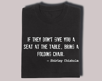 以下が含まれることがあります： 白文字で「IF THEY DON'T GIVE YOU A SEAT AT THE TABLE, BRING A FOLDING CHAIR.」と書かれた黒いTシャツ。シャーリー・チザムの引用。シャツはきれいに折りたたまれています。