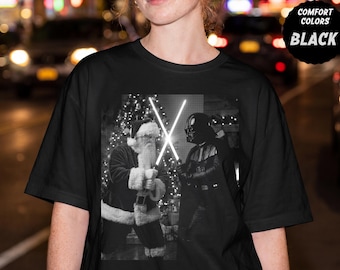 以下が含まれることがあります： 黒いTシャツで、クリスマスツリーの前で光るライトセーバーを使って決闘するサンタクロースとダース・ベイダーの白黒グラフィックプリントが施されています。右上隅には、COMFORT COLORS BLACKというテキストが入った黒い円形のバッジがあります。
