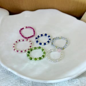 May include: Six beaded rings in various colors, including pink, white, blue, green, and gray, are displayed on a white, leaf-shaped plate. The rings feature different bead types, such as pearls and gemstones, creating a delicate and colorful jewelry collection.
