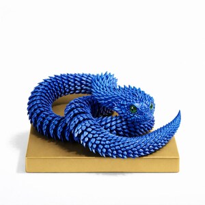 Könnte beinhalten: Detaillierte Skulptur einer blauen Schlange mit strukturierten Schuppen und grünen Augen. Die Schlange ist auf einer goldfarbenen rechteckigen Basis aufgerollt, vor einem weißen Hintergrund. Die Form der Schlange ist sehr stilisiert.